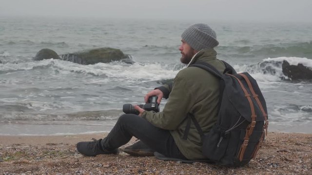bearded man taking pictures with professional camera in front of the ocean