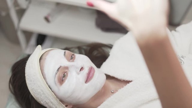 Pretty female client lying on the couch in beaty spa/salon. She is covered with white mask talking to herself, filming video, sending an air kiss, smiling