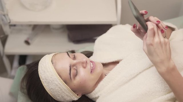Pretty female cutomer/client lying on the couch in beaty spa/salon. Woman dressed in bathing gown using her phone and smiling like a princess. Her nails are red. She is white and clean