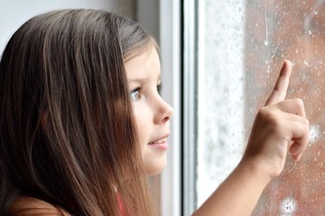 Fototapeta premium Beautiful happy toddler girl looking through the window with raindrops and drawing a finger on the wet glass. Long haired cute little girl looks at the rain outside the window. 