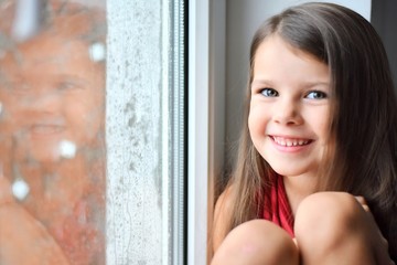 Beautiful smiling caucasian white girl sitting on a window sill and looking out the window on rainy weather. Happy young girl looked outside at raining moment. Portrait of cheerful kid at windowsill