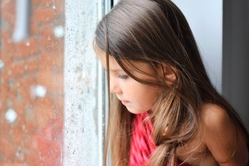 Fototapeta premium Beautiful smiling caucasian white girl sitting on a window sill and looking out the window on rainy weather. Happy young girl looked outside at raining moment. Portrait of cheerful kid at windowsill