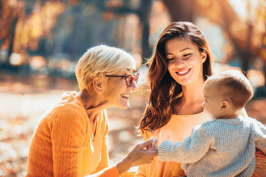 Grandmother and mother smiling at baby in autumn park.