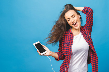 Image of happy young lady standing isolated over blue background. Using phone, listening music in headphones.