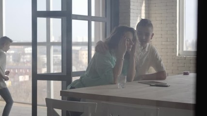 Nervous young mother trying to calm herself massaging her head at home. In the background her sons playing running around. Teen boy comes and hugs mom. Woman is tired. Difficulties in raising children