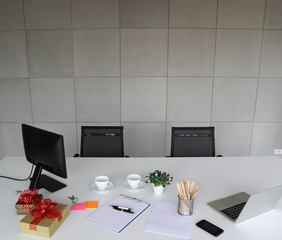 Picture of laptop, mobile phone, pens, pencils, white paper on the white desk. Also includes flower, whites cups, notepads, monitor and box of gifts. With the two black chairs.
