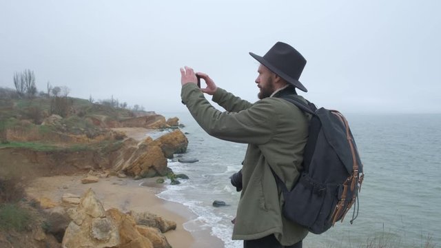 Backview guy in a hat and jacket taking pictures of beautiful natural scenes using a seaside mobile phone. Wild cold day