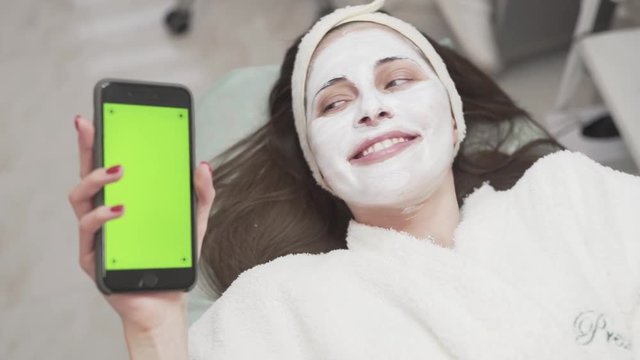 Pretty female client lying on the couch in beaty spa/salon covered with white mask, holding phone in right hand, acting playful, showing facial expression