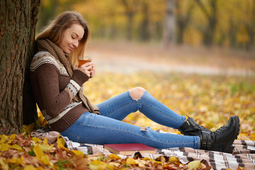 Naklejka na ściany i meble Girl in the autumn park on a warm blanket and with hot tea. Concept of autumn warmth, atmosphere and comfort
