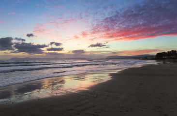 Location place Agia Marina Beach, island Crete, Greece. Sea coast spangled by rocks, the sunrise is reflecting on the wet sand. The mountains in haze on the horizon. Storm sea with high waves.