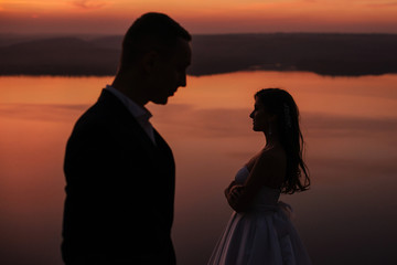 Silhouette of young wedding couple of groom and bride standing on the hill near the beautiful lake. Scenic landscape view with sunset. Bakota, Ukraine