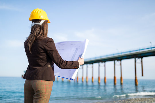 Ingegnere donna con progetto in mano vista di spalle guarda dalla spiaggia il ponte di acciaio sul mare