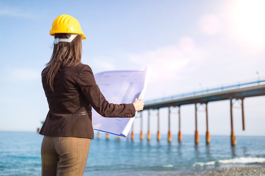 Ingegnere donna con progetto in mano vista di spalle guarda dalla spiaggia il ponte di acciaio sul mare