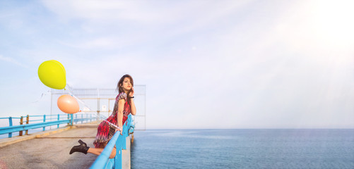 Estate. Ragazza con vestito rosso allegra su un pontile con dei palloncini colorati riempiti di elio vicino la spiaggia sul Mare.