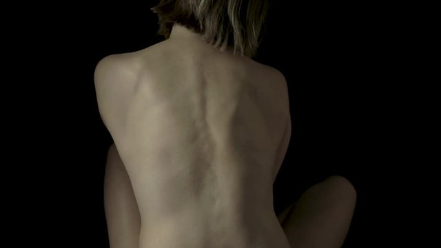 View of a naked woman sitting cross legged on a black background as she sways side to side and flexes her back muscles