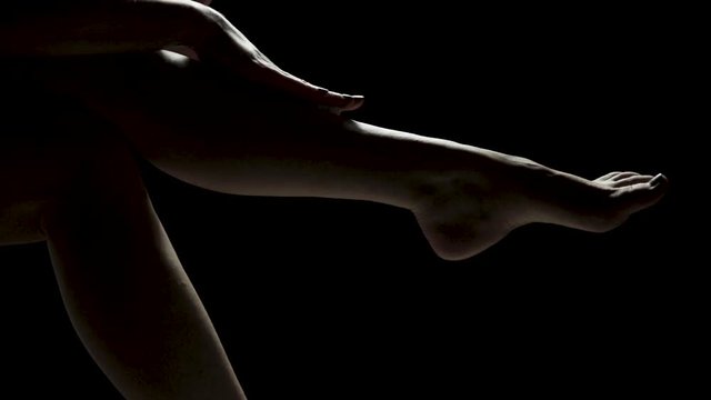 Dimly lit shot of a woman's legs as she massages lotion into her legs and feet against a black studio background