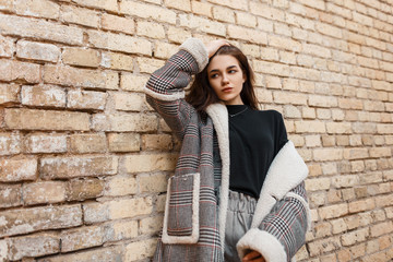 Beautiful young brunette woman in vintage gray pants in a black shirt in a fashionable plaid jacket posing standing near a brick building. Stylish girl model outdoors. Retro style.
