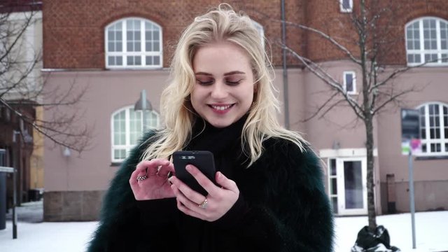 Beautiful and happy young teen girl is swiping on her phone outdoors in a cold winter time.