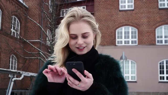 Pretty young blond teen girl is swiping on a mobile dating app, outside in a winter time.