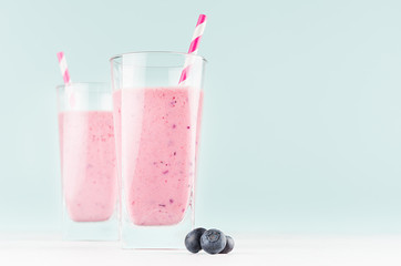 Healthy dairy dessert of ripe blueberry in two glass with straws, handful berries on white wood board and pastel green mint background, copy space.