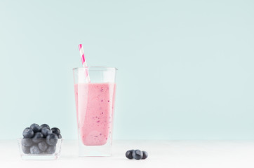Diet purple milk beverage in elegant glass with striped straw, blueberry in bowl on pastel soft light blue background, copy space.