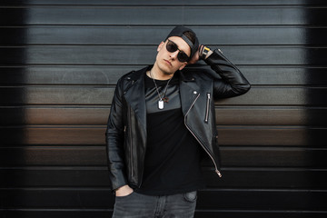 Serious young stylish hipster man in fashionable black clothes in vintage gray jeans in a cap in...