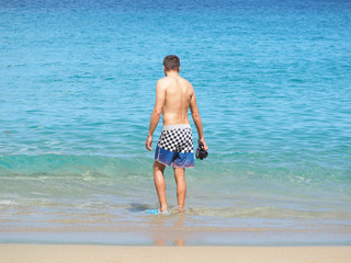 Cropped image of a man walking into the ocean, wearing shorts and flippers, holding a camera in a housing for shooting under water on the seashore. sandy beach.