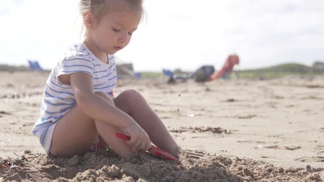 Cute little girl sitting on the sandy beach by the sea. Rebbenok buries his feet with sand. Girl holding a red shovel and digging sand