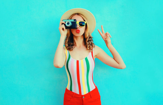 Colorful young woman holding retro camera, blowing red lips sends air kiss in summer straw hat having fun on blue wall background