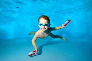 Happy little boy swimming underwater in the pool, smiling and posing for the camera with toys in his hands on a blue background. Portrait. Underwater photography. Horizontal orientation - obrazy, fototapety, plakaty