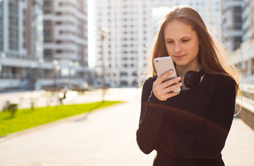 Outdoor portrait of young teenager brunette girl with long hair. girl on city in black dress looking on the smart phone walking in the street in a sunny day