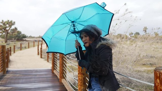 A beautiful woman in bad weather with a blue umbrella during the strong winds of a rain storm in the desert SLOW MOTION.