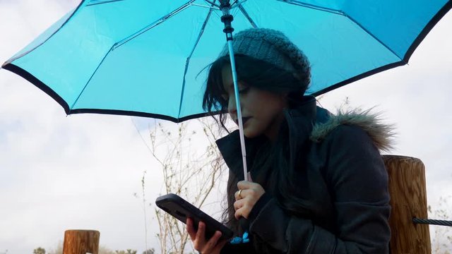Close up on a pretty woman texting on a smartphone under a blue umbrella during a windy rain storm.