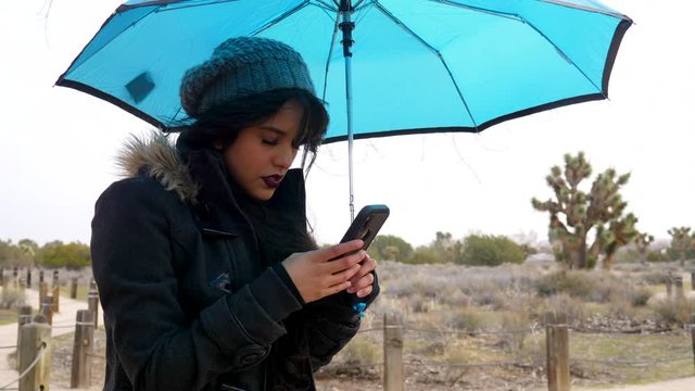 A beautiful young woman texting on her smartphone looking sad and moody during a rain storm under a blue umbrella SLOW MOTION.