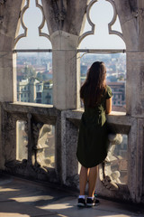 Attractive girl in a green dress and sneakers, with long dark hair, turns her back and looks out the window at the view of the city from the roof of the Milan Cathedral. Milano Duomo, Italy, observe.