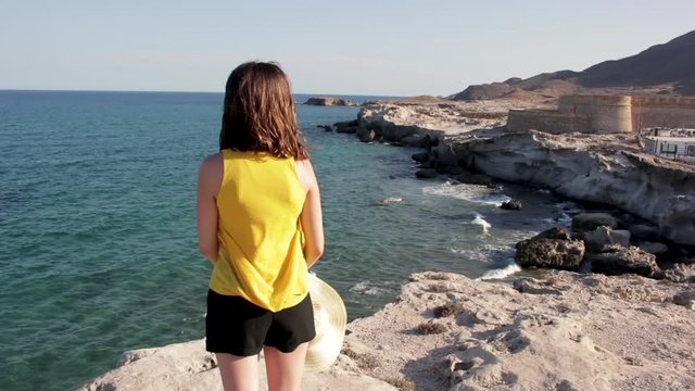 Medium shot, young girl standing on cliff, facing ocean coast Almeria, Spain