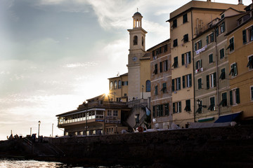 Architecture in the marine. Resedential buildings with shutter windows and tower with round clock and bell on the shore of Genoa Boccadasse, with sun rising behind the buildings.
