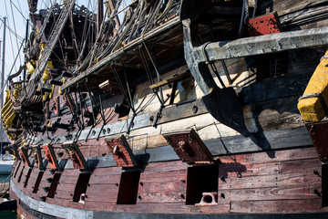 Pirate's old sailing wooden boat with opened gun compartments and canons. Sea tackles, ropes and handrails. Sunny summer day on a coast of Genoa embankment, harbour in Italy