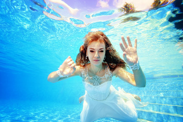 Obraz na płótnie Canvas Cute blonde bride swims underwater in the pool in a white dress and poses on camera on a Sunny day. Portrait. Close up. Wedding underwater. Underwater photography. Horizontal view