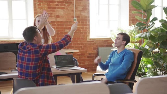 Young team of three software developers finding problem solution while discussing code on laptop computer screen and celebrating it with high fives, tracking left shot