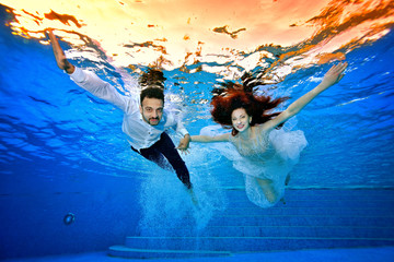 Happy bride and groom in wedding dresses swim underwater on the background of a bright sunset on the surface of the water. They smile and pose for the camera with their arms outstretched. Portrait