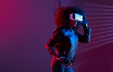 Mod curly dark haired girl dressed in black leather jacket and gloves uses the virtual reality glasses on her head in the dark studio with neon light