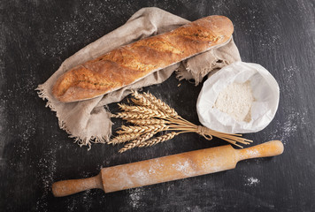 fresh bread with wheat ears, flour and kitchen utensils on dark board