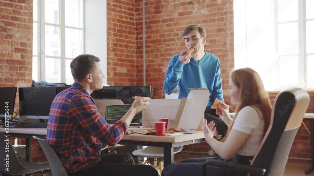Wall mural team of three young software developers sharing pizza at workplace while discussing code in office, 