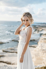 Fototapeta na wymiar Beautiful young blonde model woman with nude makeup in a fashionable wedding dress walking at the sea coast at Cyprus