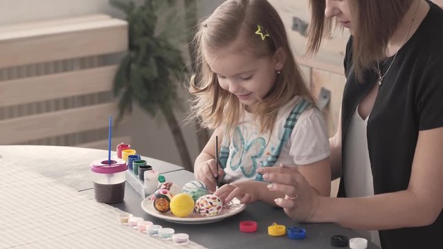 Daughter sitting on her mum's lap at table and painting the Easter eggs with gouache ushing paintbrush. Small girl touching the paintbrush and eggs.