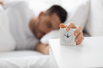 Naklejka premium morning and people concept - close up of sleepy young man in bed reaching for alarm clock on bedside table at home