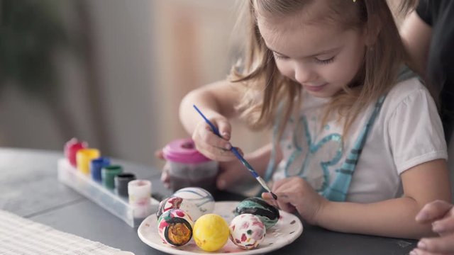 A little cute girl pains a ceramic egg in green colour with the help of brush. Her mum sts right behind her and holds a plastic can of water on the table from the right, so it does not fall.