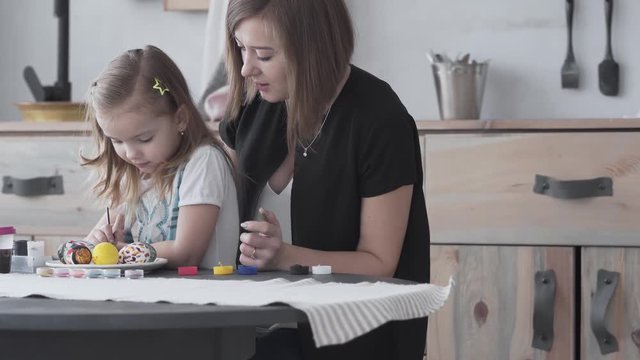 Mum together with daughter painting eggs with gouache using brush. Small girl sitting on mum's lap and mum's teaching the daughter