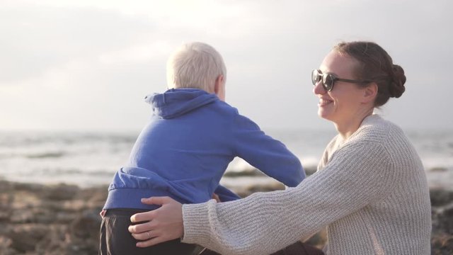 A woman sits by the sea and watches the sunset. A son comes up to her, sits down on her, they hug, and mom shows him something far into the sea with her hand